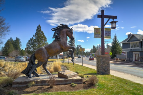 A bronze statue of a rearing horse stands near a street sign and buildings under a blue sky with clouds.