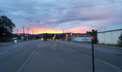 A quiet street at dusk, with a vibrant sunset behind distant mountains and buildings lining the road.