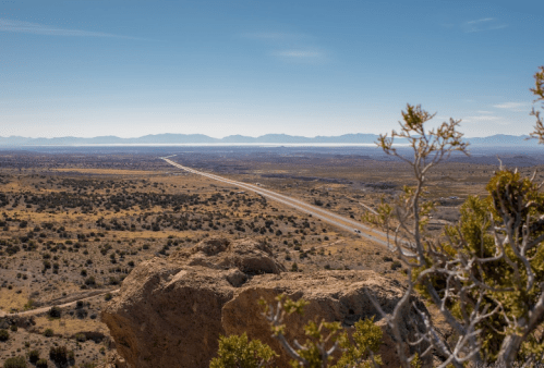 A vast desert landscape with a winding road, mountains in the distance, and sparse vegetation under a clear blue sky.