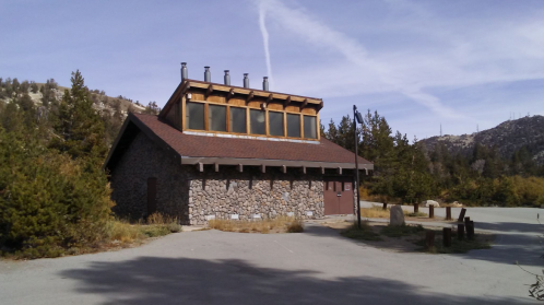 A stone building with a wooden roof, surrounded by trees and mountains under a clear blue sky.