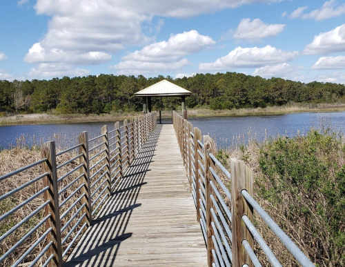 A wooden boardwalk leads to a gazebo over a calm waterway, surrounded by lush greenery and blue skies.