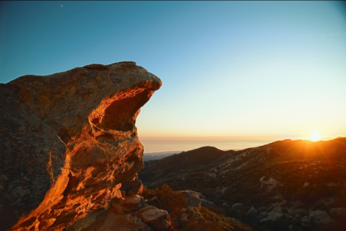 A rocky formation resembling a face overlooks a sunset over rolling hills and the ocean in the distance.