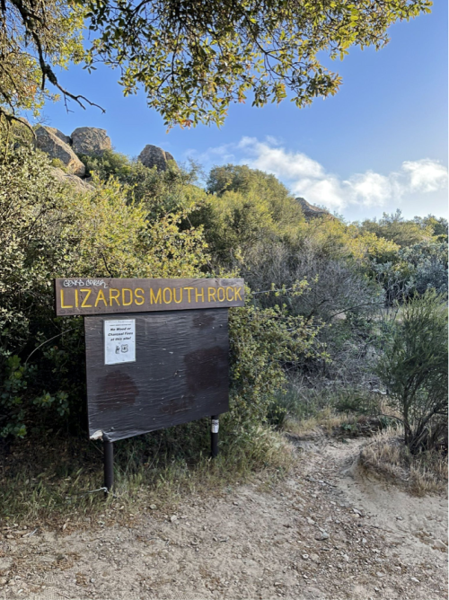 Sign for Lizards Mouth Rock along a dirt path, surrounded by greenery and rocky terrain under a blue sky.