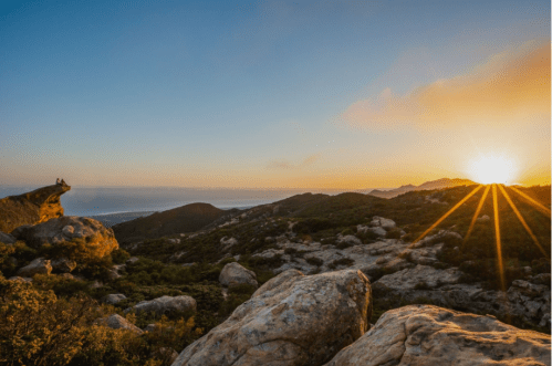 A scenic landscape at sunset, featuring rocky hills and a glowing sun over the ocean in the distance.