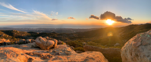 A panoramic view of a sunset over rolling hills and the ocean, with large rocks in the foreground.