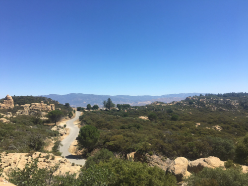 A winding road through a green landscape with rocky formations and distant mountains under a clear blue sky.