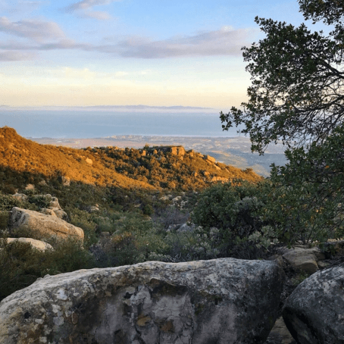 A scenic view of rolling hills and distant mountains under a partly cloudy sky, with large rocks in the foreground.