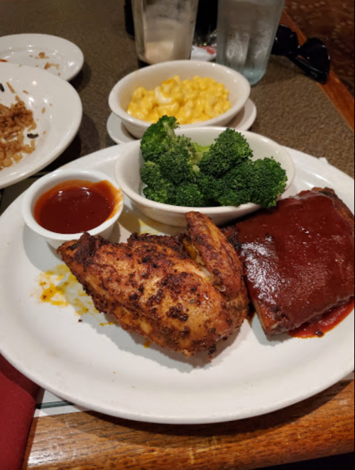 A plate with grilled chicken, barbecue ribs, macaroni and cheese, and steamed broccoli, served with dipping sauce.