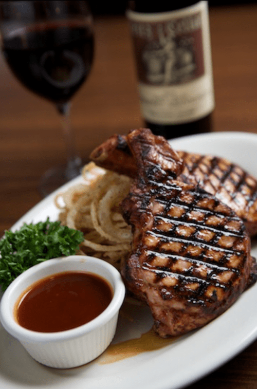 Grilled pork chops on a plate with onion rings, a side of sauce, and a glass of red wine in the background.