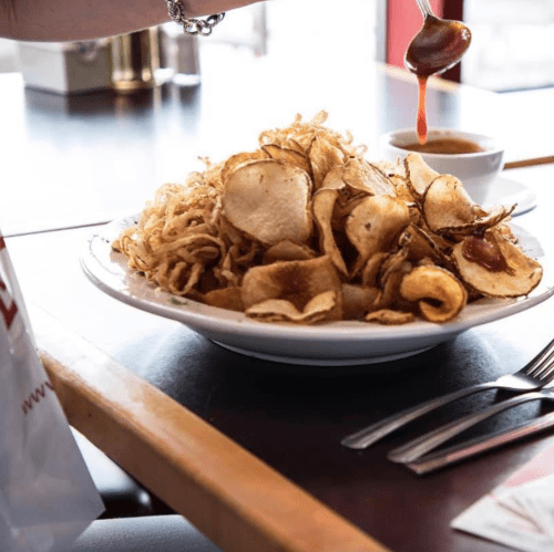 A plate of crispy potato chips with a small bowl of dipping sauce, served on a table with utensils nearby.
