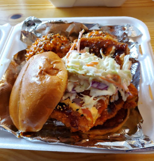 A close-up of a fried chicken sandwich topped with coleslaw, served with rice and sauce in a takeout container.