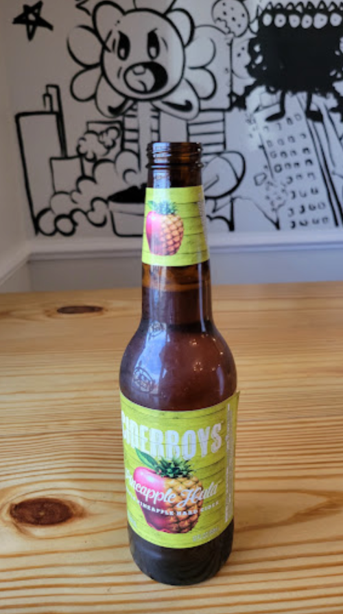 A bottle of Pineapple Hula soda on a wooden table, with a colorful mural in the background.