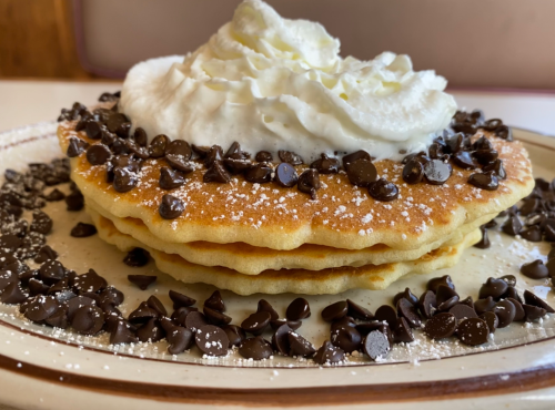 Three fluffy pancakes topped with whipped cream, chocolate chips, and a dusting of powdered sugar on a plate.