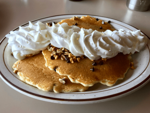 A plate of three fluffy pancakes topped with whipped cream and sprinkled with chocolate chips and nuts.