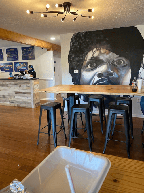 A restaurant interior featuring a large mural of a man with curly hair, wooden tables, and black stools.