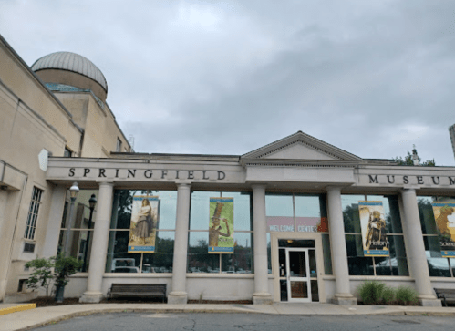Exterior of the Springfield Museum featuring large windows and a welcome center entrance under a cloudy sky.