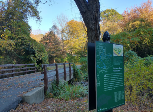 A scenic path with a wooden bridge and a sign about the Catoctin River Watershed, surrounded by autumn foliage.