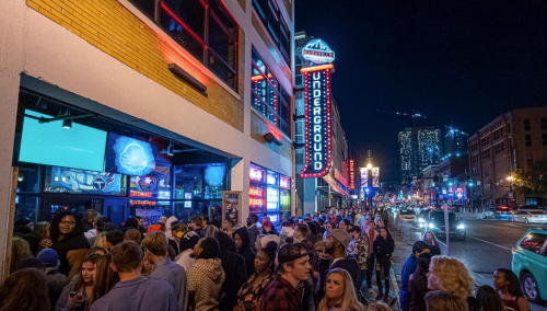 A crowded street scene at night with people lined up outside a venue, illuminated by colorful neon lights.