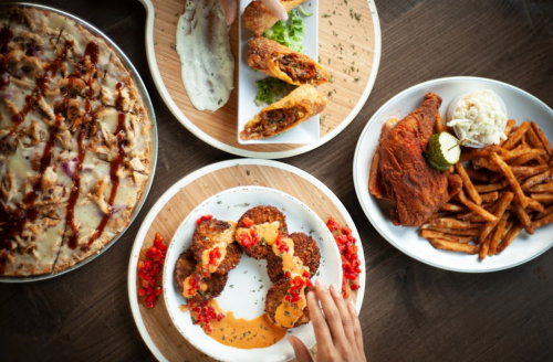 A spread of delicious dishes including pizza, fried chicken, spring rolls, and fries on wooden and white plates.