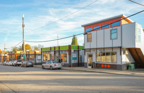 A street view of a small commercial area with colorful buildings and parked cars under a clear blue sky.