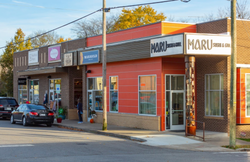 A row of shops featuring Maru Sushi & Grill, with cars parked outside and trees in the background on a clear day.