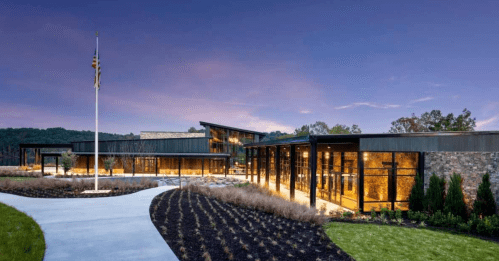 Modern building with large glass windows, surrounded by landscaped gardens and a flagpole, under a twilight sky.
