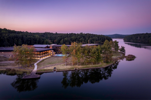A serene lakeside view at dusk, featuring a modern building surrounded by trees and calm water.
