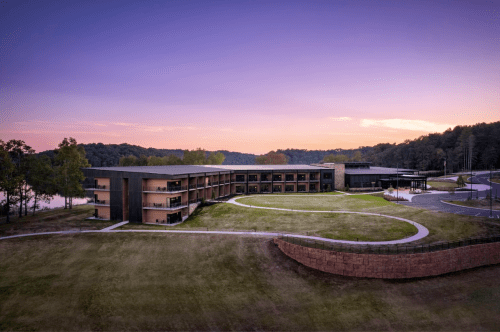 Modern building by a lake at sunset, surrounded by greenery and a winding path.
