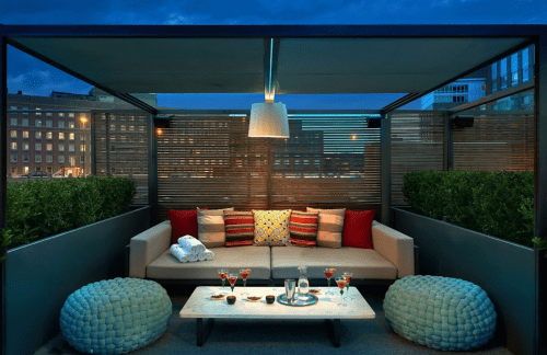 Cozy rooftop lounge with plush seating, decorative pillows, and drinks on a table, illuminated by soft lighting.