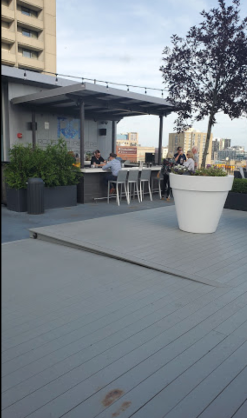 A rooftop patio with people sitting at a bar, surrounded by plants and city views in the background.