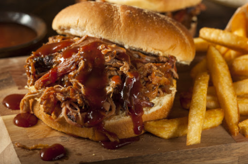 A close-up of a pulled pork sandwich topped with barbecue sauce, served with a side of golden French fries.