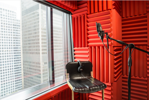 A recording studio with red soundproofing, a microphone on a stand, and a black chair by a large window overlooking a cityscape.