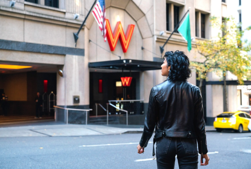 A person in a black leather jacket stands outside the W hotel, with flags and a yellow taxi in the background.