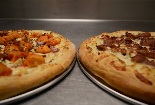 Two pizzas on metal trays: one topped with orange chunks and herbs, the other with bacon and a golden crust.