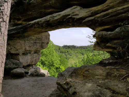A natural rock arch frames a lush green landscape with trees and a cloudy sky in the background.
