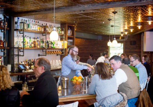A busy bar scene with patrons seated at the counter and a bartender preparing drinks in a cozy atmosphere.