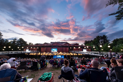 A crowd sits on the grass in front of a stage, with a colorful sunset sky and trees in the background.