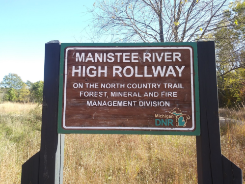 Sign for Manistee River High Rollway on the North Country Trail, managed by Michigan DNR, surrounded by grass and trees.