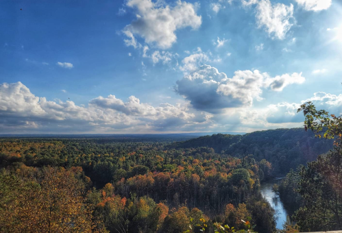 A scenic view of a river winding through a forest with autumn foliage under a blue sky with fluffy clouds.