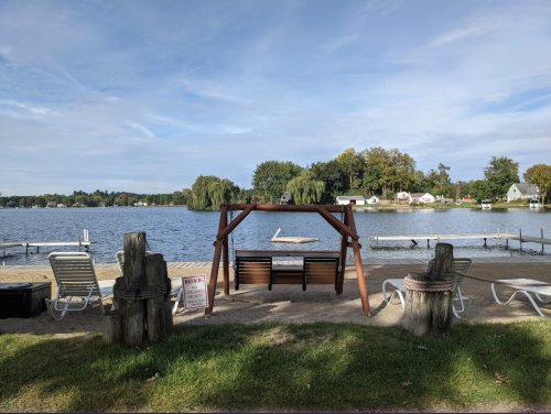 A peaceful lakeside scene with a swing set, lounge chairs, and trees along the water's edge under a blue sky.