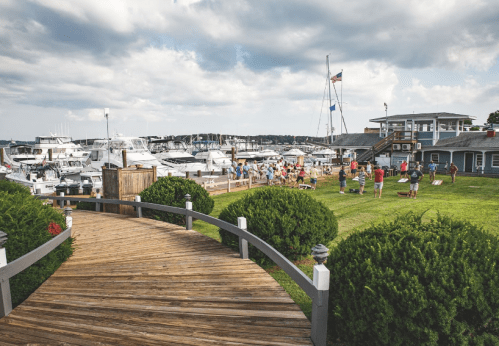 A wooden walkway leads to a marina with boats, people enjoying the outdoors, and a cloudy sky in the background.