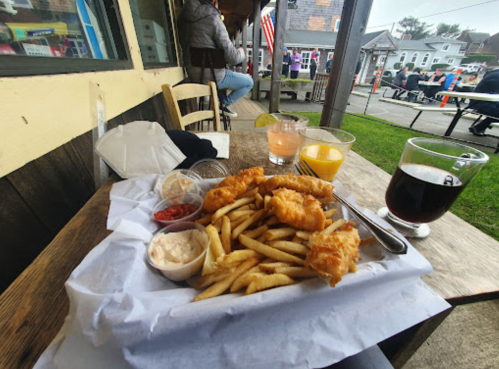 A plate of fried fish and fries with dipping sauces, accompanied by drinks, on a table outside a restaurant.