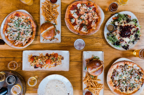 A table spread with various pizzas, burgers, fries, salads, and drinks, all arranged on wooden surfaces.