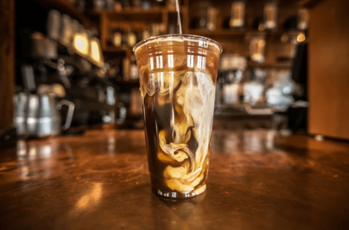 A clear cup filled with iced coffee, swirling with cream, set on a wooden table in a café.