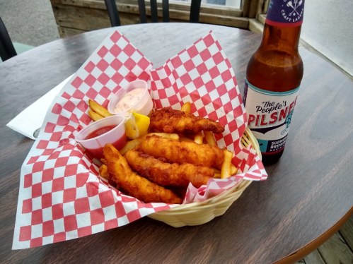 A basket of golden fried fish and fries with dipping sauces, alongside a bottle of Pilsner beer on a wooden table.