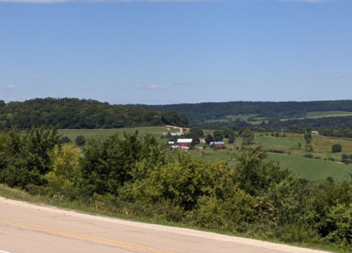A scenic view of rolling green hills and farmland under a clear blue sky, with a road in the foreground.