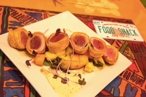 A plate of seared tuna rolls with a creamy sauce, garnished with microgreens, on a colorful patterned table.
