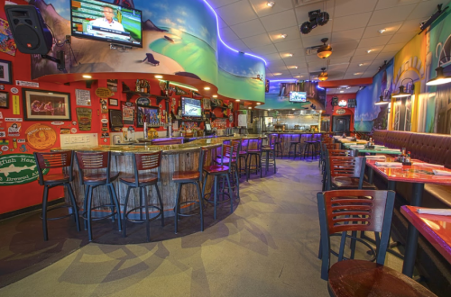 A vibrant restaurant interior with colorful murals, a bar, and tables, featuring TVs and a lively atmosphere.