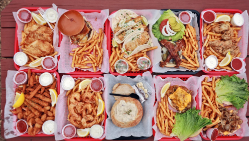 A variety of delicious meals on trays, including burgers, tacos, shrimp, and fries, with dipping sauces and lemon wedges.