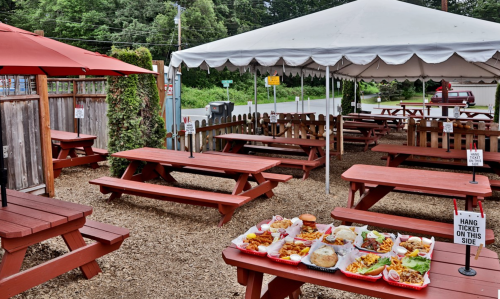 Outdoor dining area with red picnic tables, a tent, and a variety of food served on a table in the foreground.